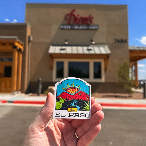 A person holds an "El Paso Sticker" from Dion's Fan Shop, showcasing a sun and mountain design with a pizza-themed sunset vibe, in front of a building displaying the "Dion's Pizza-Salad-Subs" signage.