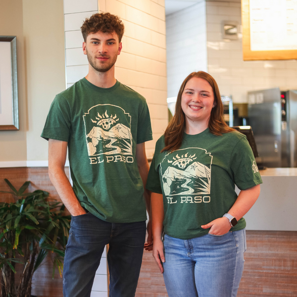 Two individuals stand indoors wearing matching "El Paso" t-shirts from Dion's Fan Shop. The man, Reyes Gonzalez, has his hands in his pockets, and the woman has her right hand in her pocket. Both are smiling.