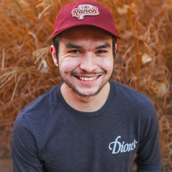 A person wearing a Dion's Fan Shop Ranch Cap in red, paired with a navy shirt accented by leather patches, smiles against a backdrop of dry grass, fully capturing the adventurous spirit of outdoor activities.