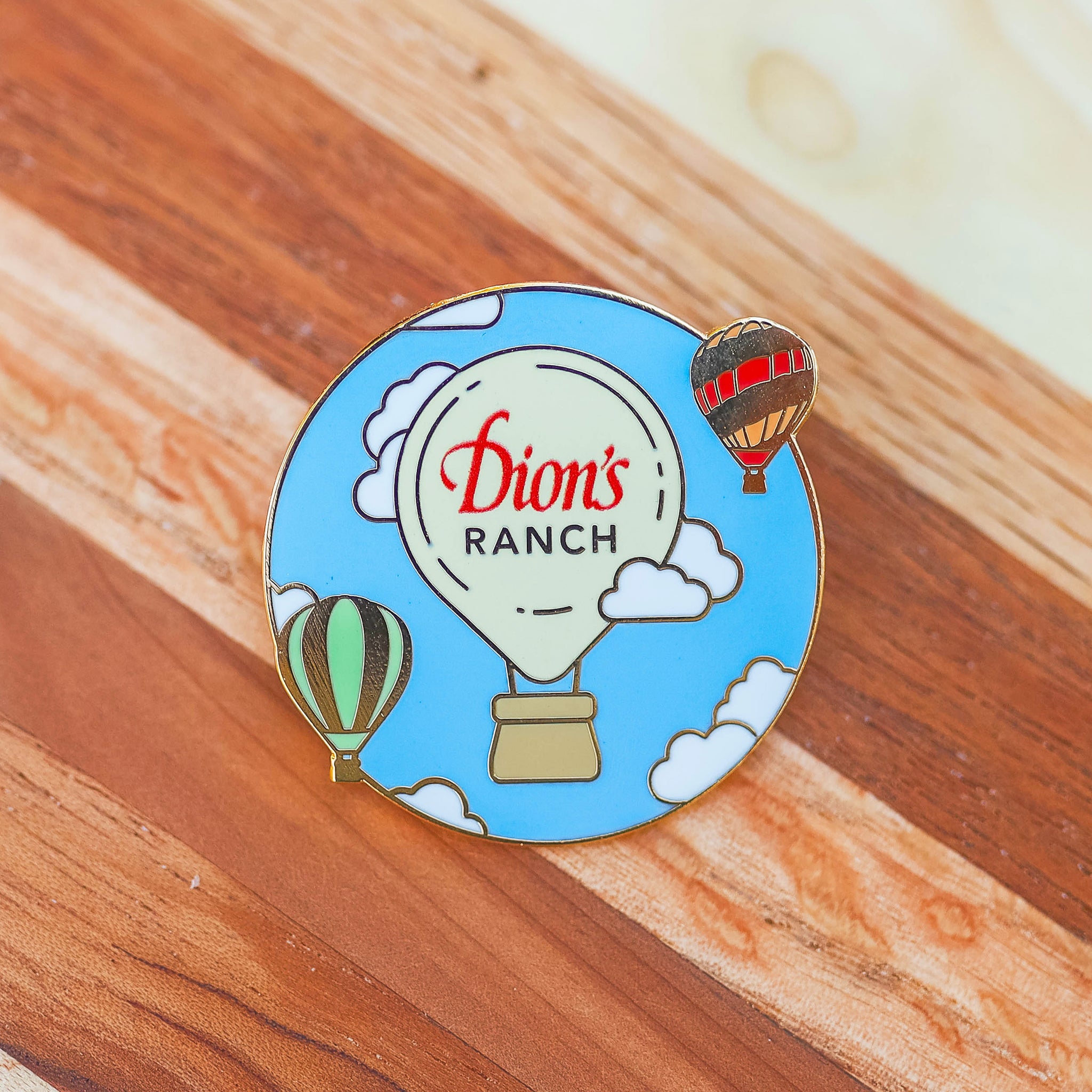 Introducing the "Dressing Up the Skies Pin - Limited Edition" from Dion's Fan Shop: a round enamel pin showcasing a hot air balloon with the text "Dion's Ranch" accompanied by two smaller hot air balloons, ideal for adorning your backpack, all set against a wooden backdrop.