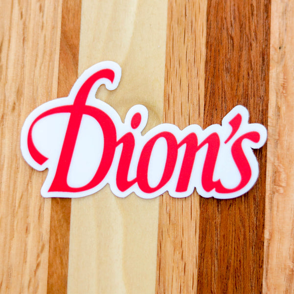 The Dion's White Sticker from Dion's Fan Shop features "Dion's" in red cursive on a white background, shown here on a striped wooden surface.