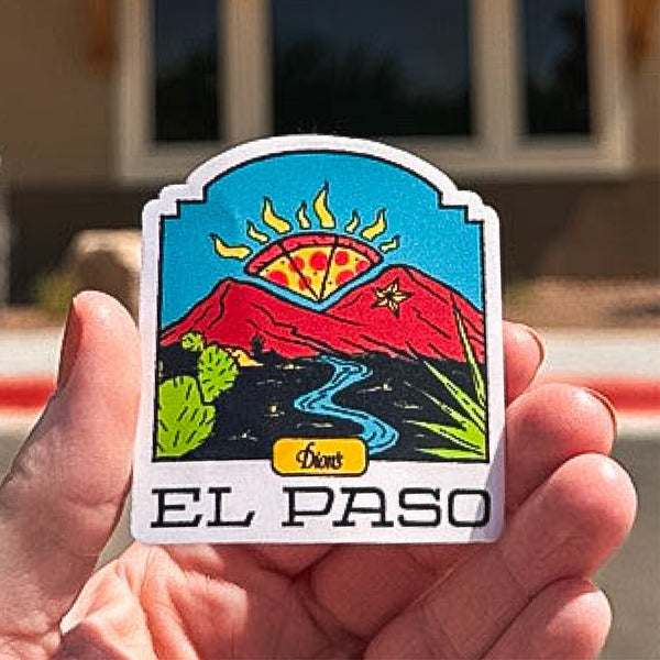 A hand holds the El Paso Sticker by Dion's Fan Shop, showing the Franklin Mountains, a winding river, cacti, and a pizza slice sun, with "El Paso" at the bottom.