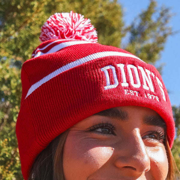 Someone wears the cozy Collegiate Beanie from Dion's Fan Shop—a red knit hat with a white pom-pom and "DION'S EST. 1978"—while standing outdoors amid green trees.