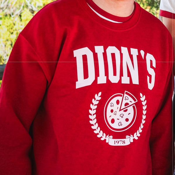 A fan wears the Dion's Fan Shop Collegiate Crewneck—red with "DION'S," a pizza graphic, "1978," and a laurel wreath—a must-have campus look.