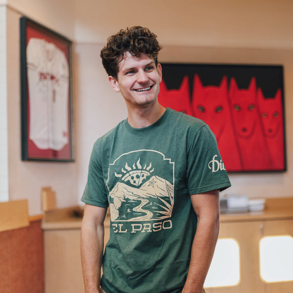 A man with curly hair stands indoors, smiling, wearing the green El Paso Tee from Dion's Fan Shop. Behind him are framed artwork, a baseball jersey, and an El Paso sticker.