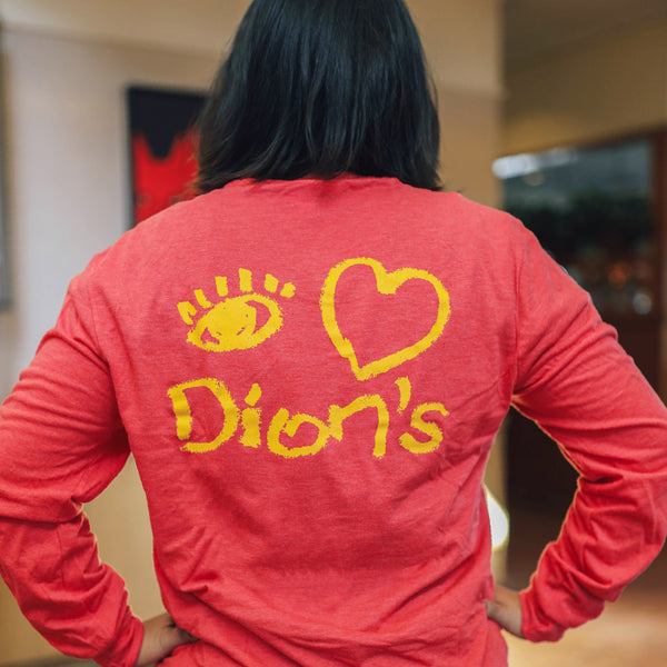 A person wears the Dion's Fan Shop "Tour - Long Sleeve Tee," a vintage red shirt featuring yellow eye, heart, and "Dion’s" graphics on the back, standing indoors with hands on hips.