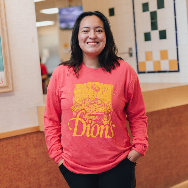 Indoors, a person smiles at the camera wearing Dion's Fan Shop's Tour - Long Sleeve Tee in vintage red with a yellow chef logo, paired with black pants.
