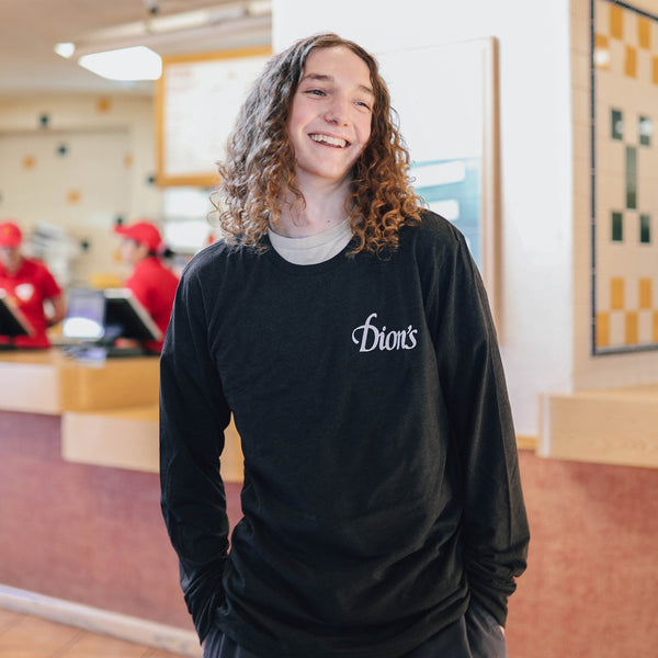 A young person with long curly hair, wearing the Dion's Fan Shop Est. 1978 - Long Sleeve Tee, stands indoors smiling with a restaurant counter and staff in the background.