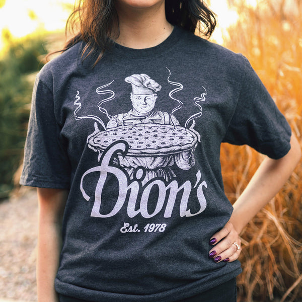 Woman wearing the Pizza Man Tee from Dion's Fan Shop—a vintage gray shirt featuring a chef with a pizza and "Est. 1978" graphic.