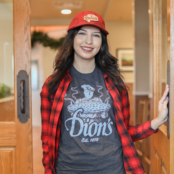 A woman in a red cap, red plaid shirt, and a vintage gray Pizza Man Tee from Dion's Fan Shop stands smiling in a doorway.
