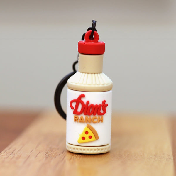The Ranch Bottle Keychain from Dion's Fan Shop features a bottle-shaped design with a pizza slice graphic, shown on a wooden surface.