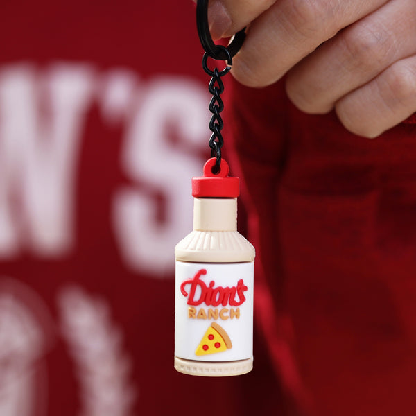 A hand holds the Ranch Bottle Keychain from Dion's Fan Shop, shaped like a bottle labeled "Dion’s Ranch" and featuring a small pizza slice graphic on the label.