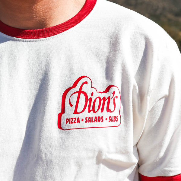 Wearing the Retro Ranch Tee from Dion's Fan Shop, featuring a white retro design with red trim and a "Dion's Pizza Salads Subs" logo on the chest for ultimate vintage vibes.