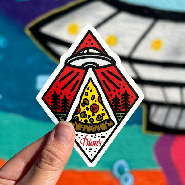 A hand holds a Roswell Dion's Sticker from Dion's Fan Shop, featuring a UFO beaming up pizza with trees and a red sky; a mural of a UFO is blurred in the background.