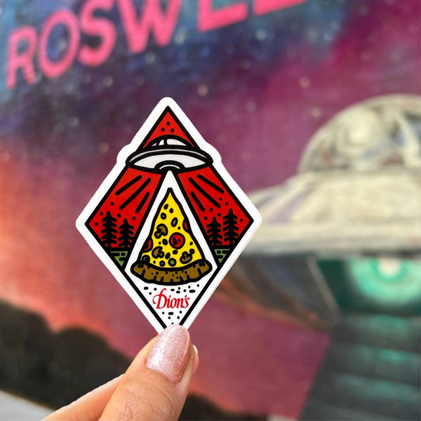 A hand holds the Roswell Dion’s Sticker from Dion’s Fan Shop, featuring a UFO beaming up pizza with “Dion’s” text; a “Roswell” mural in the background highlights it as a quirky New Mexico souvenir.