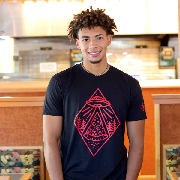 A young man with short curly hair smiles indoors, wearing the Dion's Fan Shop Roswell Tee featuring a red geometric UFO and trees graphic.