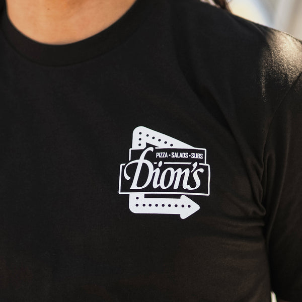 A person wears the Route 66 Centennial Tee from Dion's Fan Shop—a black shirt featuring a white "Dion's Pizza • Salads • Subs" logo, celebrating Albuquerque’s iconic flavors and the Route 66 Centennial.