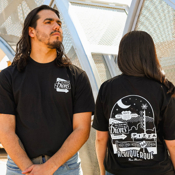 Two people wear black Route 66 Centennial Tees from Dion's Fan Shop in Albuquerque—one shows the front logo, while the other displays the back graphic.