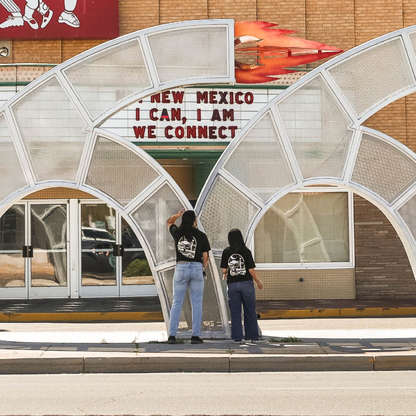 Two people in black graphic t-shirts, one wearing the Route 66 Centennial Tee from Dion's Fan Shop, stand facing a metal arch on an Albuquerque sidewalk near a building with a "NEW MEXICO I CAN, I AM, WE CONNECT" sign.