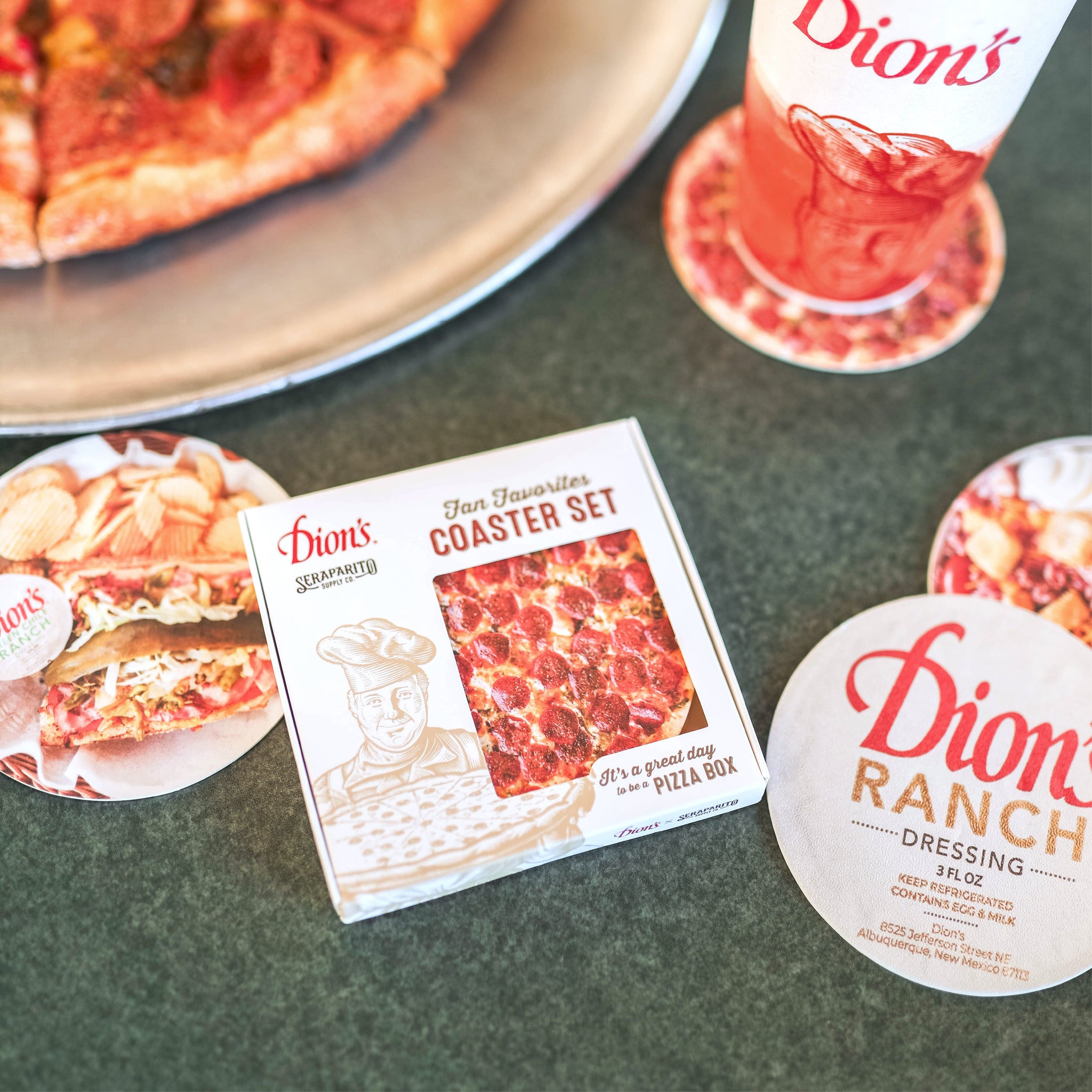 A Dion’s pizza on a metal tray sits near a Dion's Fan Shop x Seraparito Fan Favorites Coaster Set—plus a soft drink cup and branded promo items—creating a cheesy gift scene on a green tabletop.
