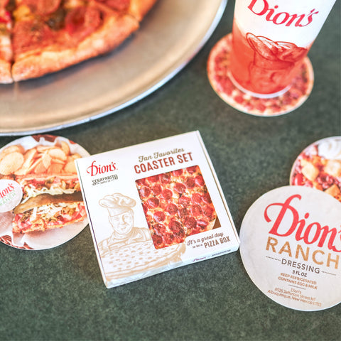 A Dion’s pizza on a metal tray sits near a Dion's Fan Shop x Seraparito Fan Favorites Coaster Set—plus a soft drink cup and branded promo items—creating a cheesy gift scene on a green tabletop.