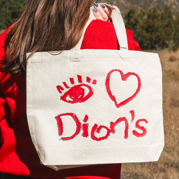A person with long brown hair holds Dion's Fan Shop’s eco-friendly Canvas Bag outdoors, featuring red artwork of an eye, a heart, and the word "Dion’s.