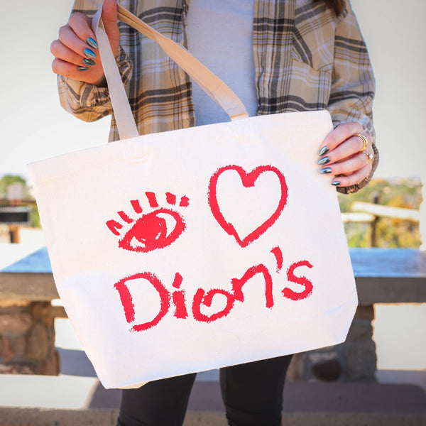 A person wearing a plaid shirt stands outside holding the Dion’s Fan Shop Canvas Bag featuring eco-friendly canvas with red "I [eye symbol] [heart] Dion’s" text and drawings.