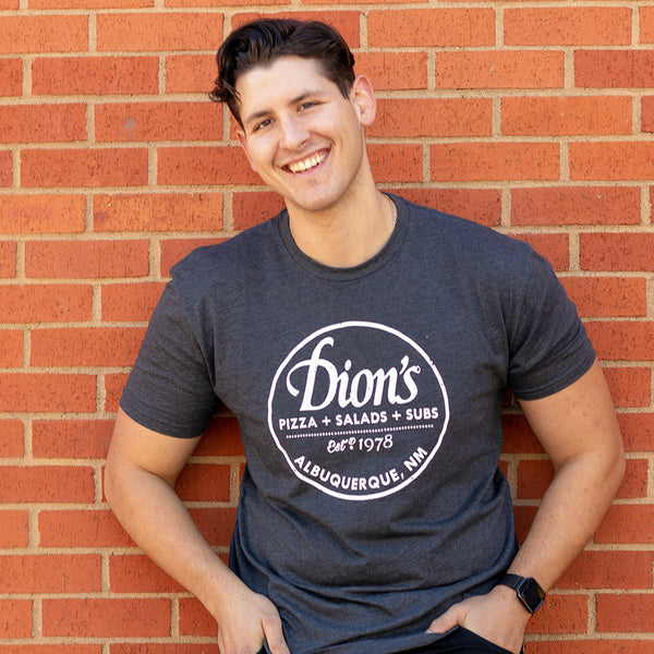 Wearing the Est. 1978 Tee from Dion’s Fan Shop, a person leans against a red brick wall, smiling with one hand in their pocket.