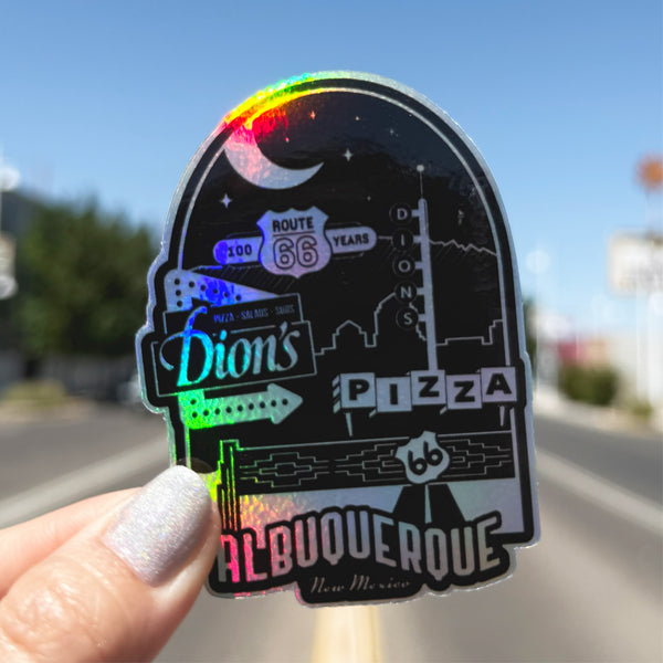A hand holds the Route 66 Centennial Sticker from Dion's Fan Shop, featuring Dion's Pizza and Albuquerque, NM, with a blurred street scene in the background.