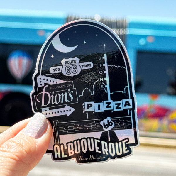A hand holds a Dion's Fan Shop Route 66 Centennial Sticker, featuring Albuquerque, New Mexico, with a blue bus and hot air balloon blurred in the background.