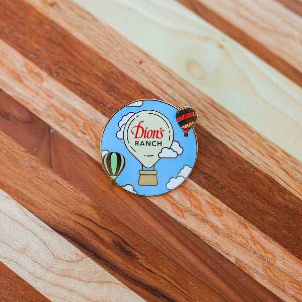 Introducing the "Dressing Up the Skies Pin - Limited Edition" from Dion's Fan Shop: a round enamel pin showcasing a hot air balloon with the text "Dion's Ranch" accompanied by two smaller hot air balloons, ideal for adorning your backpack, all set against a wooden backdrop.