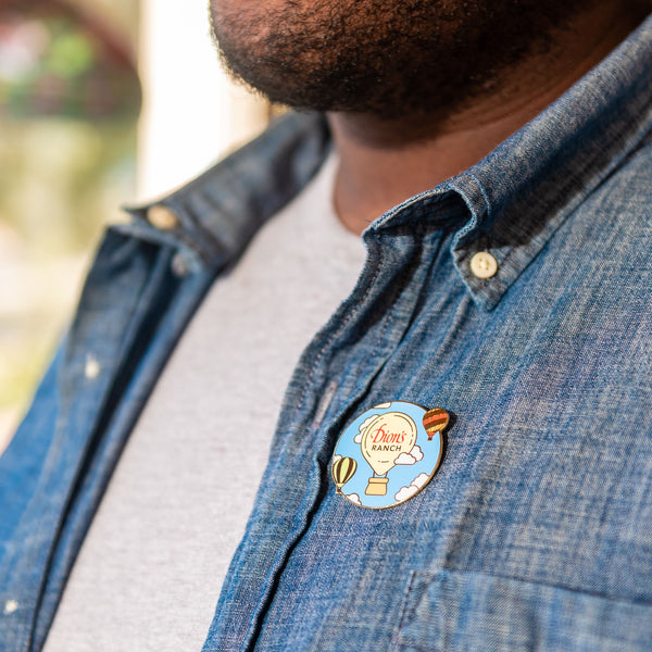 A person wearing a denim shirt with the "Dressing Up the Skies Pin - Limited Edition" from Dion's Fan Shop, featuring an image of a hot air balloon and the words "Dixon's Ranch," on the chest.