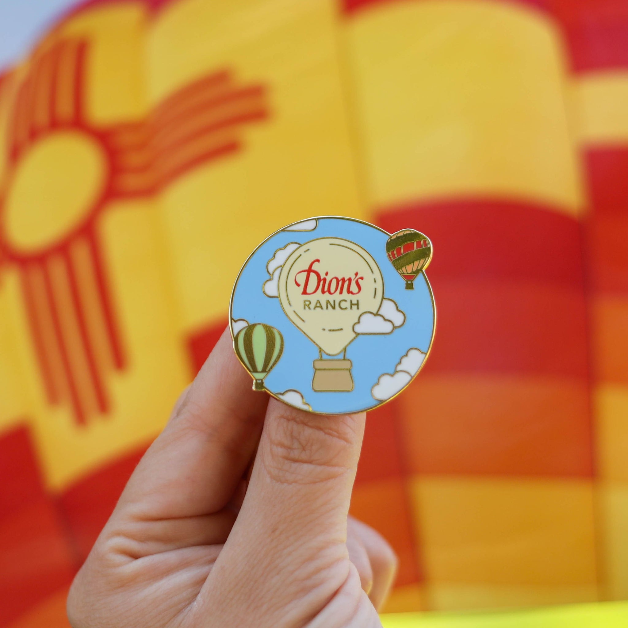 A hand holds the Dressing Up the Skies Pin - Limited Edition Pin from Dion's Fan Shop, which features three hot air balloons. A large, yellow and red hot air balloon fills the background, making this the perfect backpack accessory.