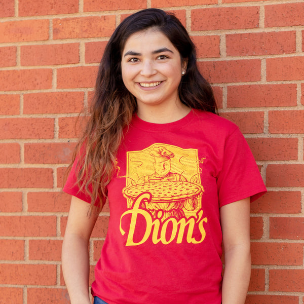 A woman with long brown hair smiles in front of a brick wall, wearing the Tour Tee from Dion's Fan Shop, looking ready for a fun field trip.