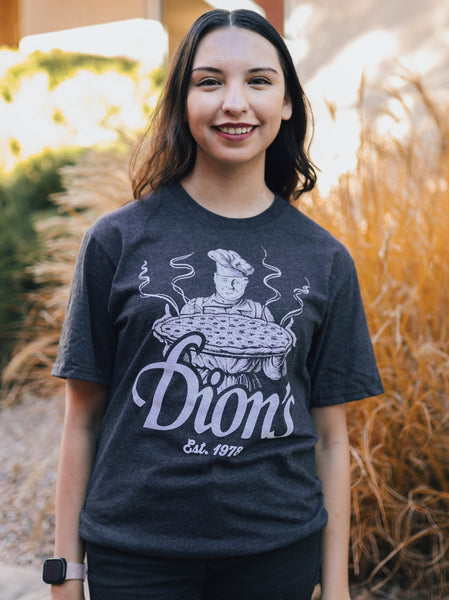 A person smiling, wearing the Pizza Man Tee from Dion's Fan Shop, a premium vintage gray T-shirt featuring a graphic of a chef holding pizza with the text "Dion's Pizza Est. 1978.