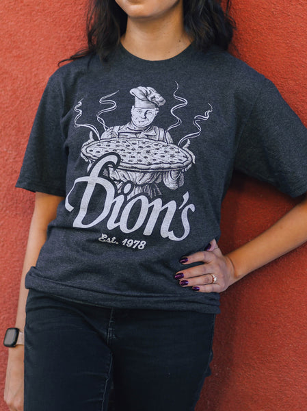 A person wearing a vintage gray "Pizza Man Tee" from Dion's Fan Shop, showcasing a chef holding a pizza, stands against a red wall.